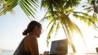 Woman with laptop working beachside under palm tree