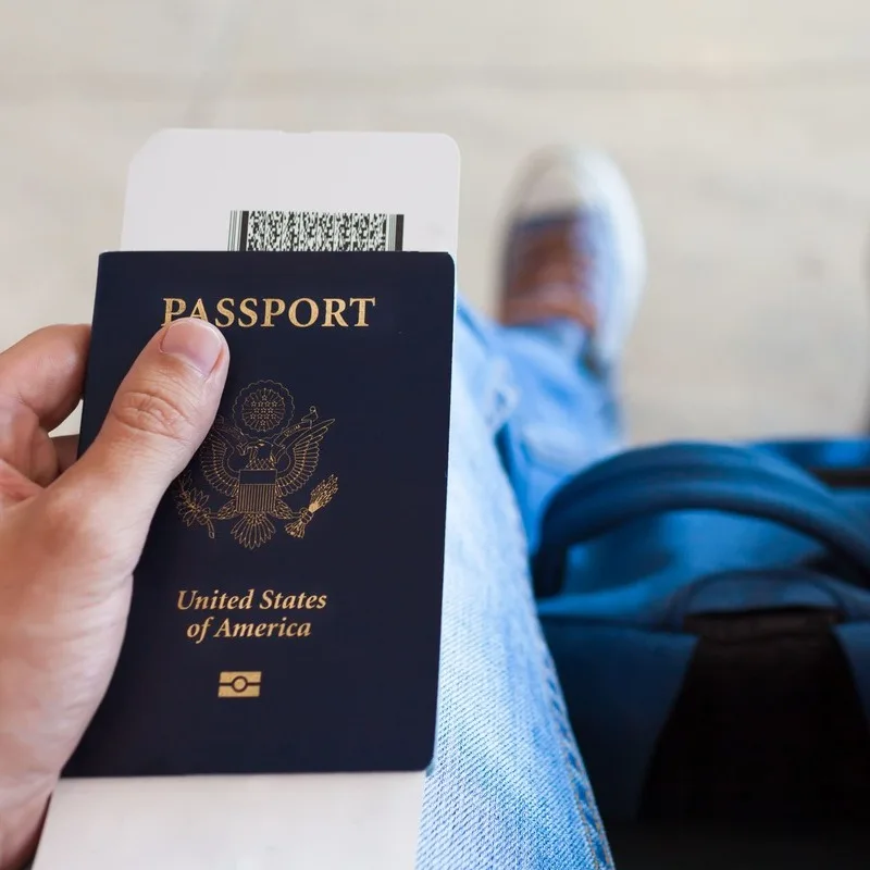 Young Male Traveler Holding Up A US Passport And A Boarding Card At The Airport